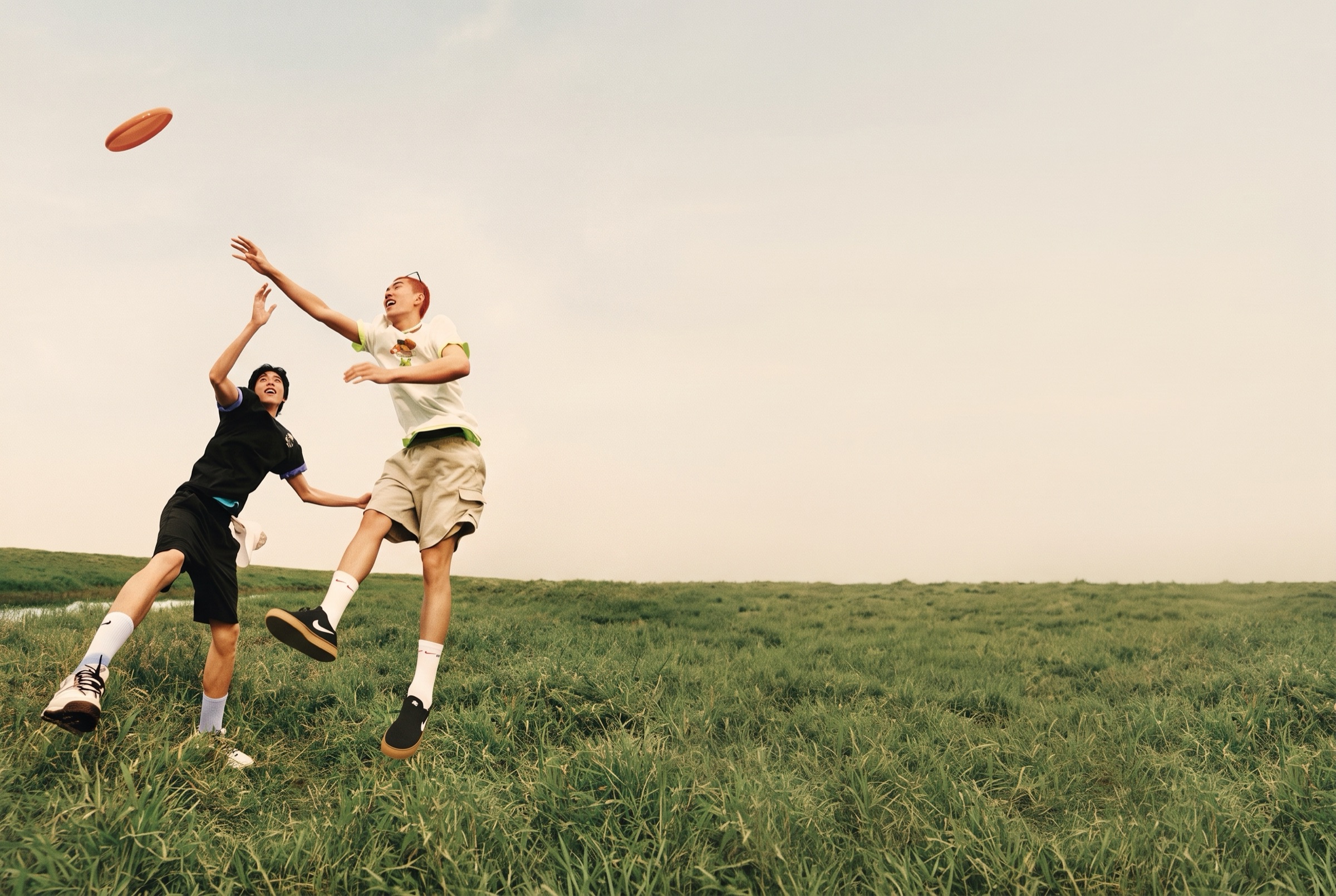 Two friends leaping for a frisbee on an open grass field under a hazy warm sky